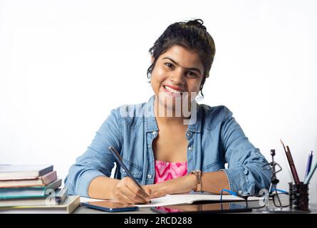 Una giovane studentessa indiana che studia e guarda la macchina fotografica sul tavolo dello studio e sullo sfondo bianco Foto Stock