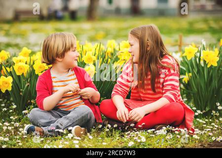 Due bambini carini, il bambino e la sorella maggiore, che giocano nel parco tra i fiori di narcisi gialli, indossando abiti rossi accesi Foto Stock