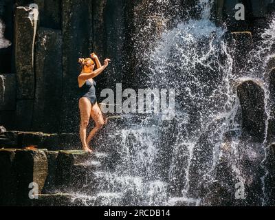 Donna caucasica in un costume da bagno nero rinfrescante sotto la caduta di corsi d'acqua che scorrono su cascate di pietra vulcanica nera. Cascate di Rochester - popul Foto Stock