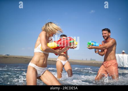 Tre giovani amici si divertono in acqua giocando con le pistole ad acqua. Amici in vacanza. Divertimento, insieme, vacanza, stile di vita. Foto Stock