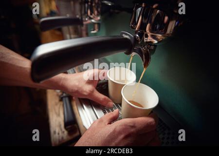 vista dall'alto delle mani maschili caucasiche che tengono due tazze di carta sotto il getto di caffè espresso nero Foto Stock