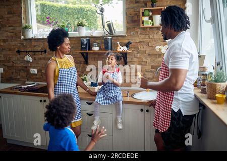 La famiglia afro-americana si riunisce intorno al bancone della cucina. La madre, il padre, il figlio e la figlia sono riuniti, con la bambina seduta Foto Stock