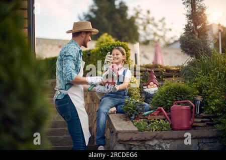 Padre e sua figlia lavorano insieme in giardino. Il padre guida con attenzione la sua bambina mentre lei apprende impazientemente del giardinaggio. Foto Stock