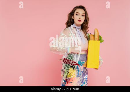 concetto di casalinga, bella giovane donna che tiene una borsa in rete riutilizzabile con generi alimentari, moglie elegante che fa i compiti di casa tutti i giorni, in piedi su sfondo rosa, Foto Stock