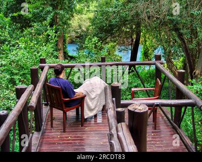 donna anziana che si rilassa su una sedia di legno sulla terrazza vicino al fiume in un villaggio di pensionati Foto Stock