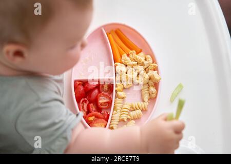 Il bambino carino mangia cibo sano pasta e verdure cotte al vapore,. Ritratti di una bella bambina di 10 mesi. Il bambino è seduto su un seggiolone speciale per Foto Stock