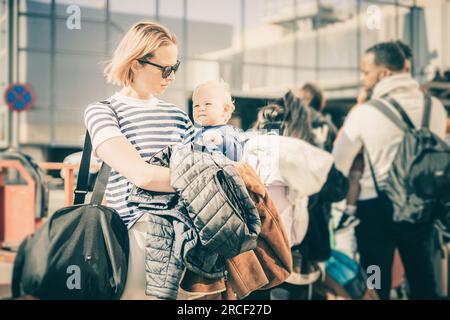 La madre viaggia con il suo bambino. Mamma che tiene in mano la borsa da viaggio e il suo bambino piccolo mentre fa la fila per l'autobus di fronte all'aeroporto Foto Stock
