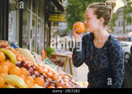 Vista laterale di una giovane-adulta felice che prende la frutta con la mano, odora un'arancia quando fa shopping in un mercato alimentare e sceglie cosa comprare al piatto Foto Stock