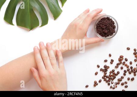 Le mani delle donne e lo scrub del caffè sulla pelle. Il concetto di cosmetici naturali. Composizione piatta di scrub di caffè, mani, chicchi di caffè e monstera Foto Stock