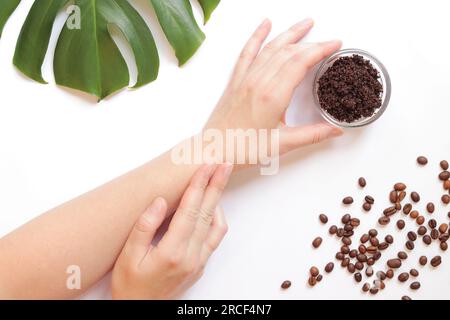 Le mani delle donne e lo scrub del caffè sulla pelle. Il concetto di cosmetici naturali. Composizione piatta di scrub di caffè, mani, chicchi di caffè e monstera Foto Stock