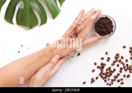 Le mani femminili applicano uno scrub al caffè sulla pelle. Il concetto di cosmetici naturali. Composizione piatta di scrub di caffè, mani, chicchi di caffè e monstera Foto Stock