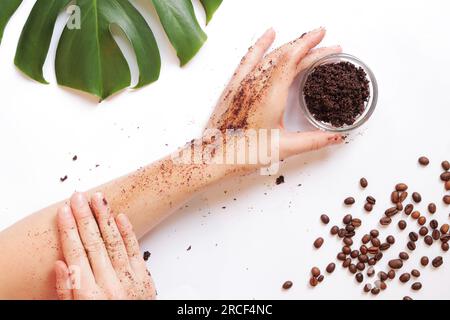 Le mani femminili applicano uno scrub al caffè sulla pelle. Il concetto di cosmetici naturali. Composizione piatta di scrub di caffè, mani, chicchi di caffè e monstera Foto Stock