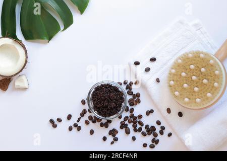 Concetto di cura personale della spa. Composizione piatta di scrub per caffè, spazzola massaggiante, asciugamano bianco, chicchi di caffè, cocco e foglia di monstera su sfondo bianco. Foto Stock