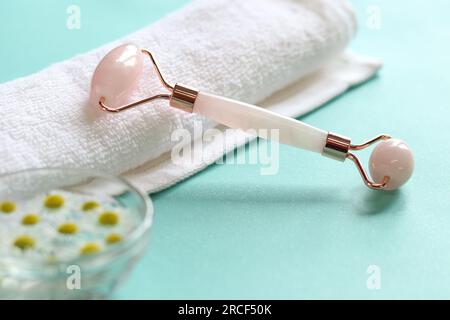 Rullo facciale in quarzo rosa su asciugamano arrotolato con sfondo blu. Rullo per massaggio viso cosmetico e acqua con fiori di camomilla Foto Stock