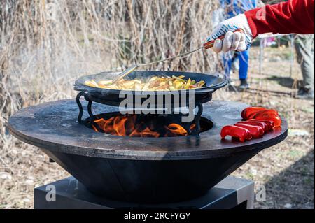 Le verdure sono tostate su un focolare rotondo. Griglia rotonda a forma di ciotola con un fuoco all'interno. L'uomo prepara il focolare per il barbecue. La legna da ardere sta bruciando Foto Stock