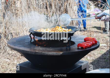 Le verdure sono tostate su un focolare rotondo. Griglia rotonda a forma di ciotola con un fuoco all'interno. L'uomo prepara il focolare per il barbecue. La legna da ardere sta bruciando Foto Stock