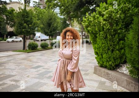 La giovane e bella donna afro-americana ama passeggiare nel parco verde pubblico estivo. Una donna felice in taglia più grande che indossa un abito rosa di seta sorride per le strade della città Foto Stock