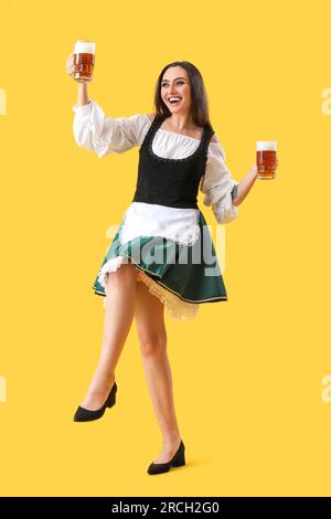 Bellissima cameriera dell'Octoberfest con birra che balla su sfondo giallo Foto Stock