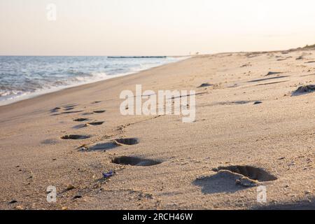 Uno scenario paesaggistico di onde che si infrangono sulla spiaggia, case sullo sfondo, conchiglie e impronte nella sabbia, cuore d'amore scritto nella sabbia. Foto Stock