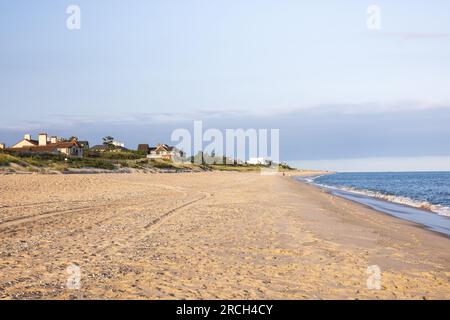 Uno scenario paesaggistico di onde che si infrangono sulla spiaggia, case sullo sfondo, conchiglie e impronte nella sabbia, cuore d'amore scritto nella sabbia. Foto Stock