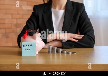 Donna in abito da lavoro seduta al tavolo con salvadanaio, pile di monete e testo PRESTITO STUDENTESCO su biglietto adesivo Foto Stock