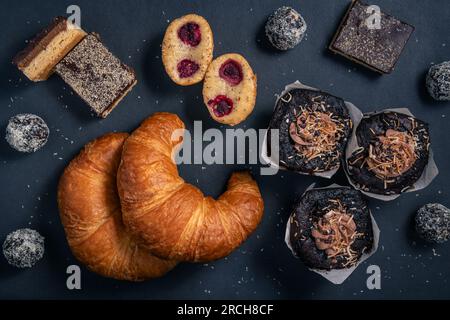 Prodotti da forno su sfondo scuro. Fette di caramello, croissant, muffin, palline proteiche, patatine fritte ai lamponi, catturato dall'alto, vista dall'alto, posizionamento piatto. Foto Stock