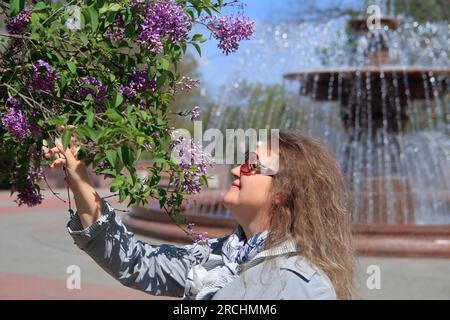 La foto è stata scattata nella città Ucraina di Odessa. Nella foto, una donna attraente con gli occhiali da sole gode dell'odore di lilacs fioriti in una città p Foto Stock