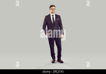 Giovane uomo caucasico con occhiali e sorriso in abito da lavoro si erge su sfondo grigio Foto Stock