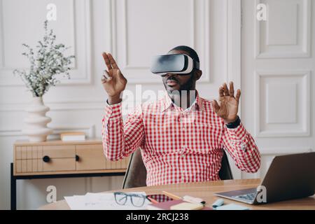 Uomo d'affari afro-americano focalizzato, visore VR acceso, immerso in un gioco di realtà virtuale; gesti, interazione con oggetti del mondo cibernetico. Foto Stock