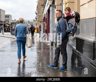 13 luglio 2023. Inverness, Highlands e Isole, Scozia. Questo è un uomo che porta il suo bambino sonnolento in uno zaino e si ripara dalla pioggia. Foto Stock