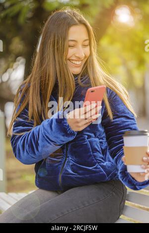 Ragazza latina che scatta una foto con il cellulare di una tazza di caffè usa e getta in un parco pubblico. Foto Stock