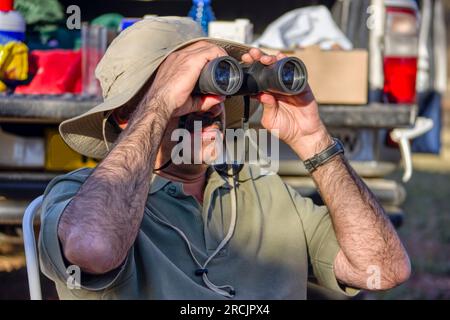 uomo arabo con il binocolo e il cappello da safari seduto su una sedia di fronte al suo veicolo 4x4 carico a guardare gli uccelli Foto Stock