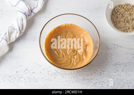 Ciotola di vetro con crema di latte condensato bollito e formaggio cremoso, e ciotola con noci macinate su sfondo azzurro, vista dall'alto. Cucinare un delizioso hom Foto Stock