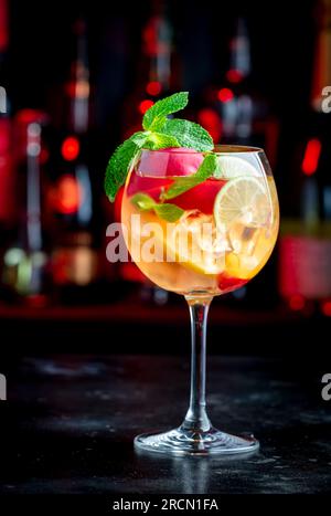 Sangria rosa bevanda alcolica estiva con vino rosa, pesca, arancia, lime e ghiaccio. Sfondo nero bar, utensili da bar in acciaio e bottiglie Foto Stock