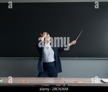 Donna caucasica dai capelli rossi con un vestito da pantaloni. Insegnante premurosa con un puntatore alla lavagna. Foto Stock