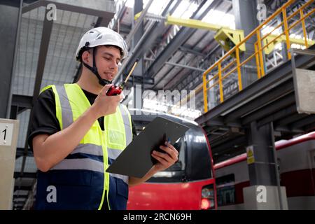 Un giovane ingegnere caucasico o un lavoratore tiene un appunti per cercare e controllare il treno elettrico per pianificare la manutenzione e utilizzare la radio per il comando Foto Stock