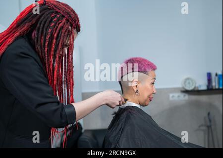 Il parrucchiere rasa il tempio di una cliente femminile. Donna asiatica con capelli corti rosa in barbiere. Foto Stock