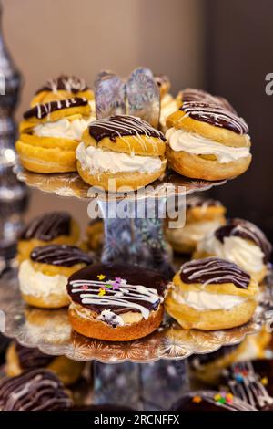 profiterole e eclairs con crema pulita e glassato con cioccolato e fondente, festa di compleanno per bambini, su una scintillante torre espositiva posta su un tavolo Foto Stock