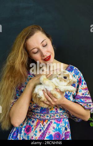 Bella donna russa con lunghi capelli biondi che tiene in mano un animale domestico di coniglio contro il muro nero Foto Stock