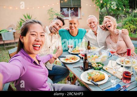 Gruppo di amici adulti adulti che sorridono scattando un selfie durante una festa al barbecue nel cortile di casa. Persone di mezza età che scattano foto sedute al tavolo del patio con espressioni positive e amichevoli. Foto di alta qualità Foto Stock
