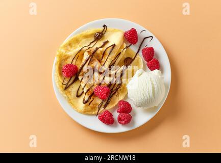 crepe appena sfornata con gelato e lamponi su piatto bianco decorato con salsa al cioccolato fuso, vista dall'alto Foto Stock