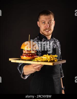 Un maestro chef sorridente in uniforme che guarda la macchina fotografica, tenendo il piatto con hamburger servito. Foto Stock