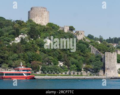 Fortezza di Rumeli circondata da alberi su una collina sulle rive del Mare del Bosforo con un traghetto passeggeri rosso attraccato, Istanbul, Turchia Foto Stock