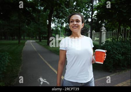 Bellissima giovane donna latinoamericana che tiene un caffè da asporto in una tazza di carta ecologica, sorridendo guardando da parte mentre cammina lungo il vicolo di un parco cittadino su una Foto Stock