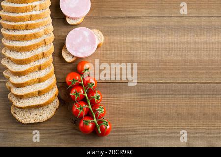 I panini con salsicce bollite si trovano su uno sfondo di legno. Pomodoro rosso ciliegia e pane bianco tagliato. Delizioso spuntino Foto Stock