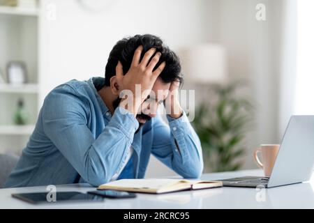 Problemi di lavoro. Indiano freelancer ragazzo seduto alla scrivania, toccando testa nella disperazione Foto Stock