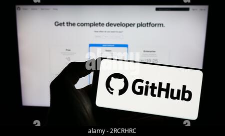 Persona che possiede il cellulare con il logo della società statunitense di sviluppo software GitHub Inc. Sullo schermo davanti alla pagina Web aziendale. Concentrarsi sul display del telefono. Foto Stock