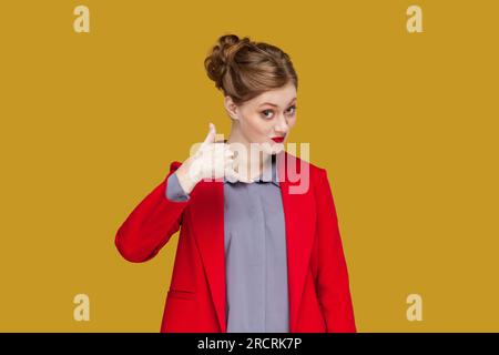 Ritratto di una donna positiva flirtante con labbra rosse in piedi che mostra il gesto di richiamami, guardando la macchina fotografica, indossando una giacca rossa. Riprese in studio in interni isolate su sfondo giallo. Foto Stock