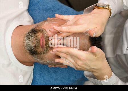 Uomo barbuto durante la procedura di pulizia del viso in una clinica di cosmetologia. Cura della pelle Foto Stock