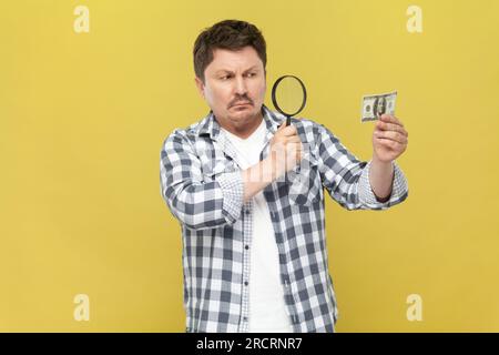 Ritratto di un uomo serio e concentrato di mezza età in camicia casual a scacchi che guarda attraverso il loupe al dollaro, verificando l'autenticità della banconota. Riprese in studio in interni isolate su sfondo giallo. Foto Stock